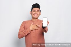 Muslim man in kufi hat smiling and presenting blank screen on mobile phone with hand on chest 0veJ7b