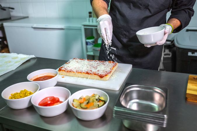 Anonymous male chef sprinkling grated cheese over a tomato sauced pizza in restaurant kitchen