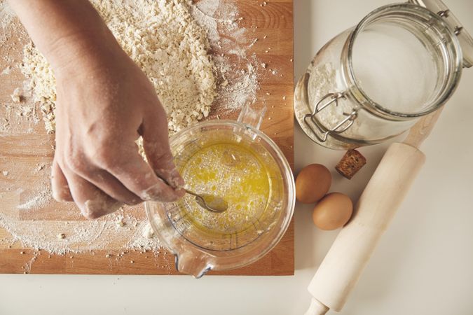 Above view of hand string oil and water in measuring cup, with dough, rolling pin and eggs