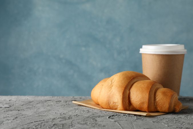 Paper cup of coffee and croissant on grey table, space for text