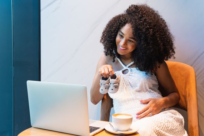 Smiling pregnant Black woman showing baby booties to laptop