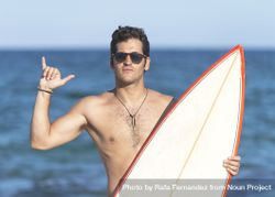 Male surfer standing with red outlined board near ocean making the “hang loose” sign 4dVVl0