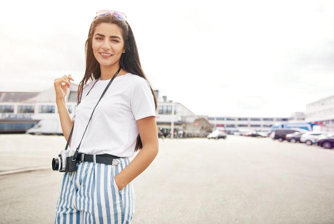 Long haired brunette woman standing outside with hand in pocket and camera around neck
