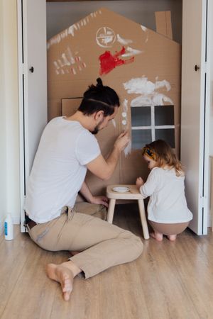 Back view of father and young daughter building a small house out of cardboard at home
