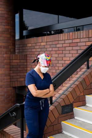 Nurse in medical PPE stands outside hospital with arms crossed and looking down
