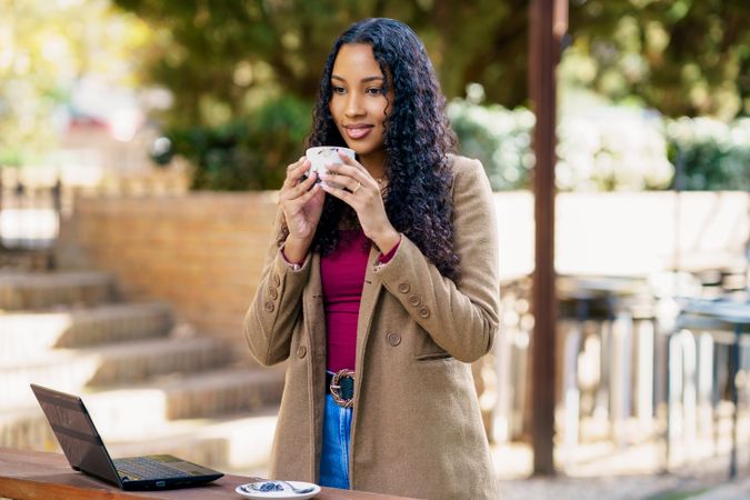 A young woman is savoring a delicious cup of coffee while sitting outdoors beside her laptop