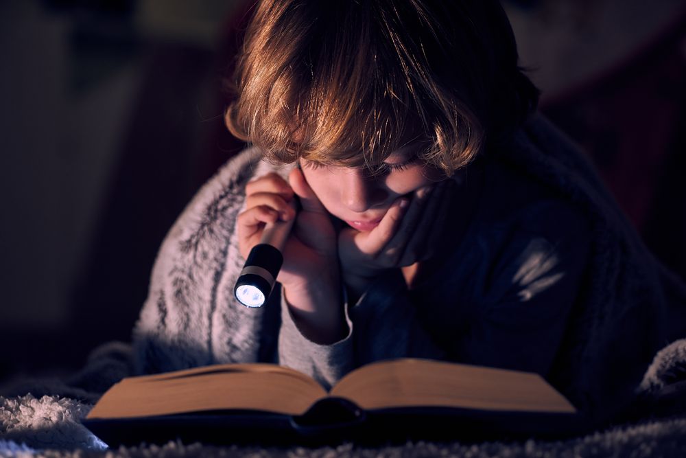 Focused boy reading book with flashlight