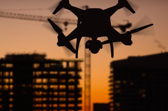 Silhouette of Unmanned Aircraft System (UAV) Quadcopter Drone In The Air Over Buildings Under Construction.