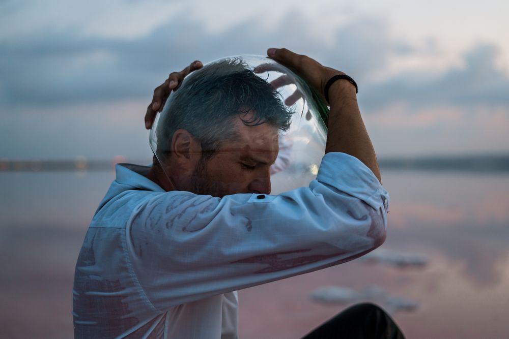 Man with fish tank on head sitting at seashore