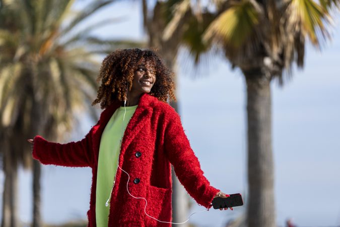 Happy female in bright colors listening to music outside on street with palm trees