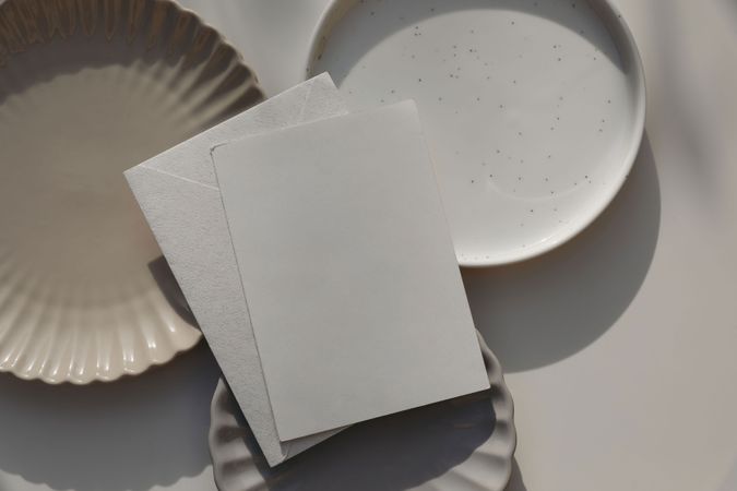 Blank greeting card, invitation mockup, envelope in sunlight. Different plates on beige table background. Soft shadows. Neutral still life, web banner. Food, restaurant, craft concept. Flat lay, top.