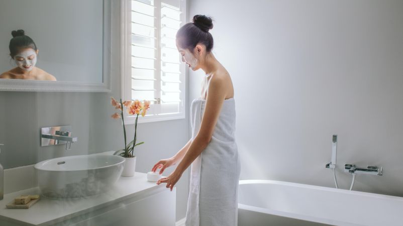 Woman wrapped in towel standing in bathroom applying moisturizing cream