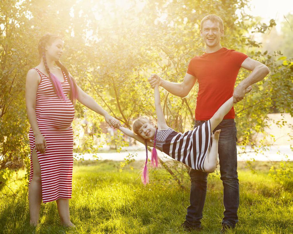 Happy expecting family of three playing in sunny park under trees