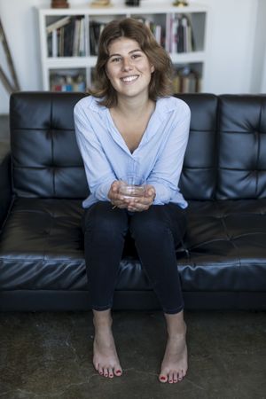 Attractive smiling young woman leaning forward on couch and holding glass of water