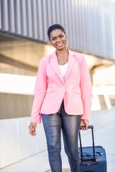 Smiling Black woman with travel bag wearing pink jacket walking outside