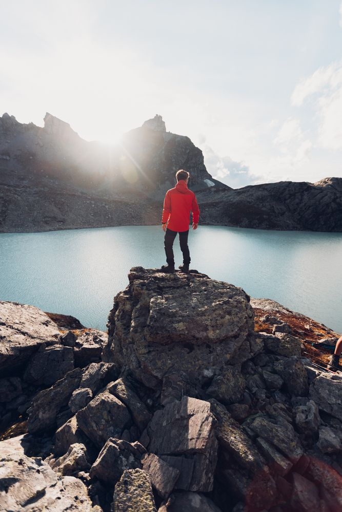 Back view of man in red jacket standing on rock by lake waterside