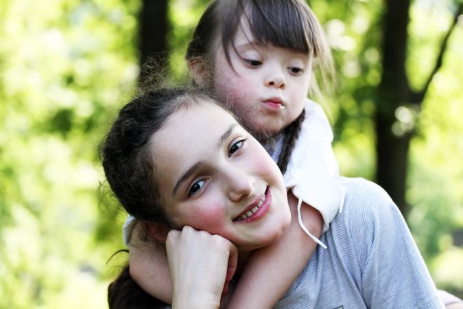 A young girl with Down syndrome hanging onto her sister’s back in the park