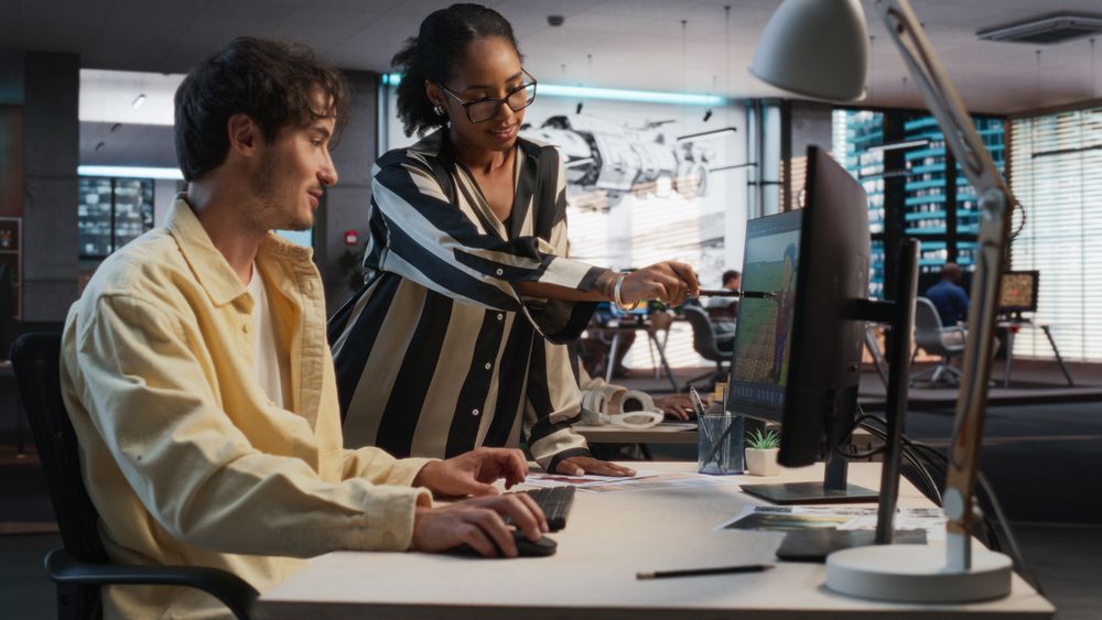 Male Game Designer Using Desktop Computer With 3D Modelling Software And Talking to Black Female Project Manager in Game Development Studio Office.