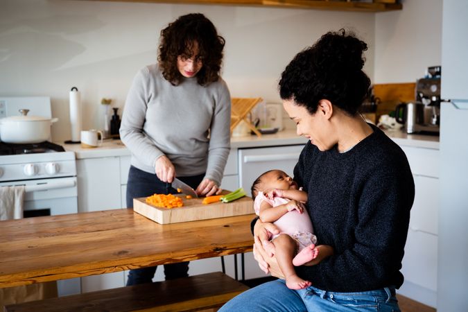 Woman holding baby while her partner chops a carrot in the kitchen