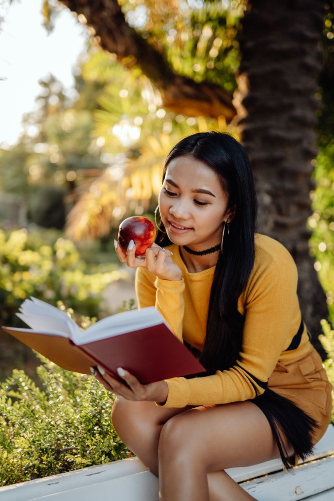 Graceful long haired Asian woman eating apple and reading book on bench in park