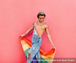 Male in open overalls looking down and holding a pride flag while leaning against a pink wall 43Wgx4