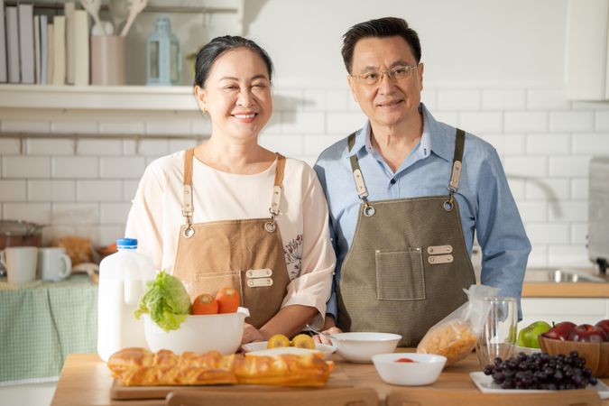 Portrait of Asian mature couple in apron cooking in the kitchen together and looking at camera