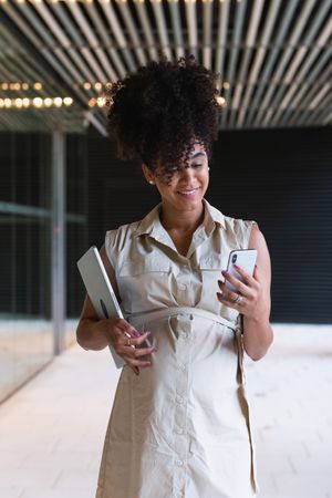 Smiling Black pregnant woman using smartphone and carrying laptop