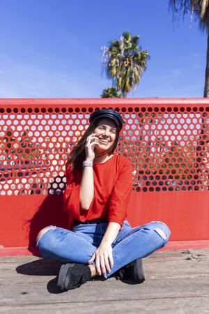 Smiling woman in red sitting cross legged talking on mobile phone outdoors on a bright day