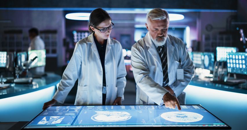 Female Neuroscientist And Male Surgeon Talking, Using Interactive Touch Screen Table With CT Scan Of Human Brain With Tumor, Finding Treatment