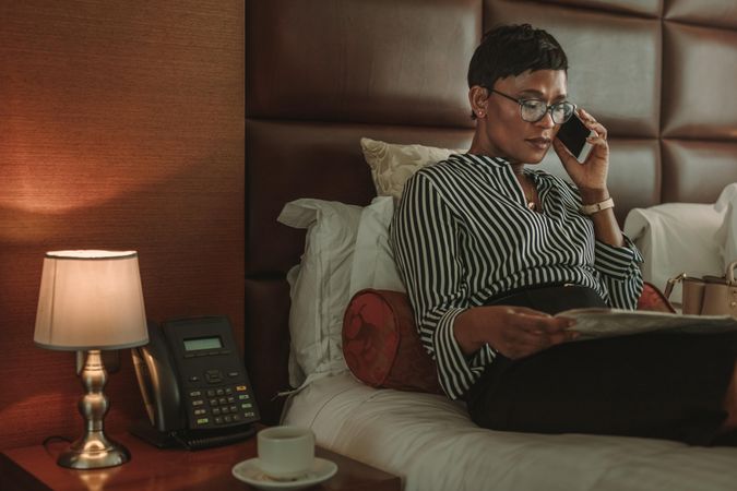 Businesswoman on hotel room bed reading newspaper and talking on cell phone