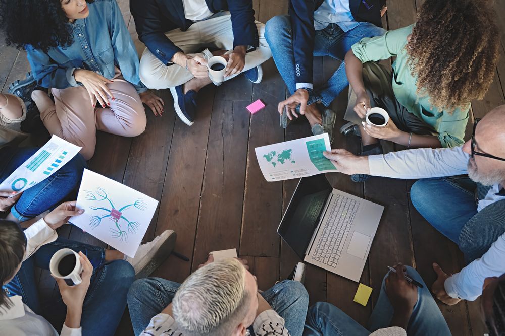 Aerial view of colleagues sitting on wooden floor during a brainstorming session, sharing coffee, laptops, and documents with charts and mind maps