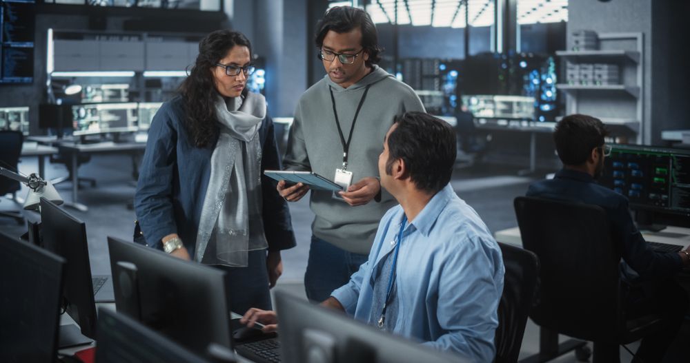 Team of Three Young Indian Software Engineers Use Computer to Discuss a Technological Project in Modern Industrial Office.