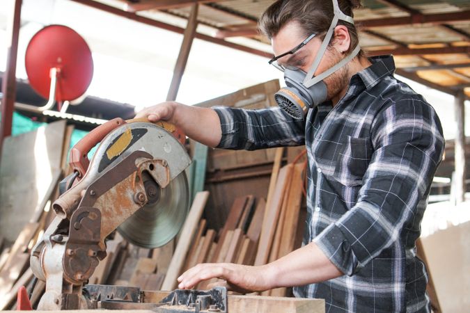 Male carpenter wearing protective mask and glasses using electric circular saw cutting wood board at the carpentry workshop