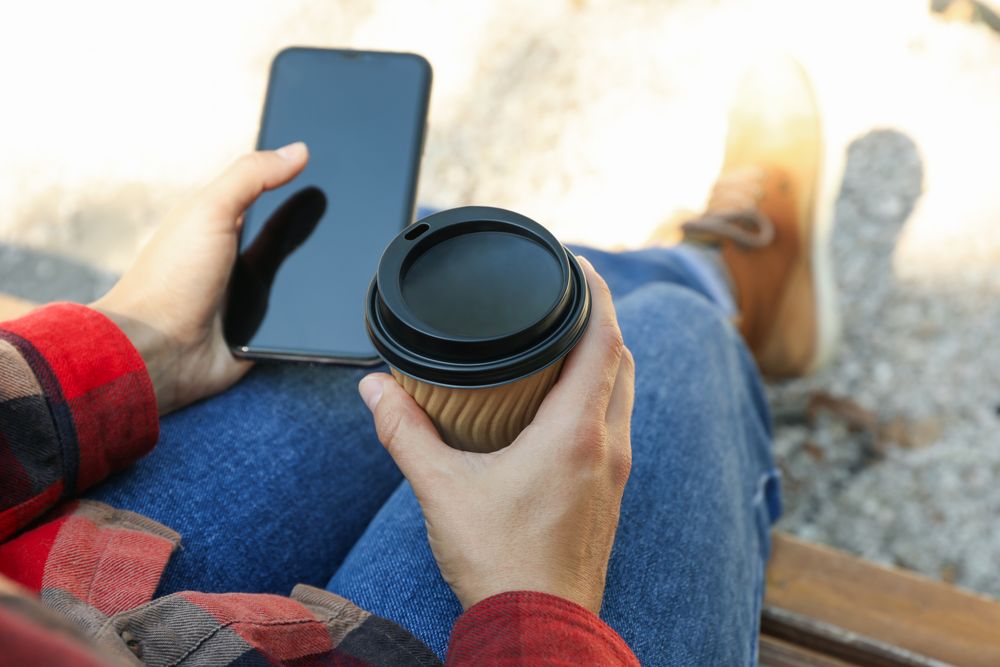 Woman in jeans and shirt hold paper cup and phone outdoor