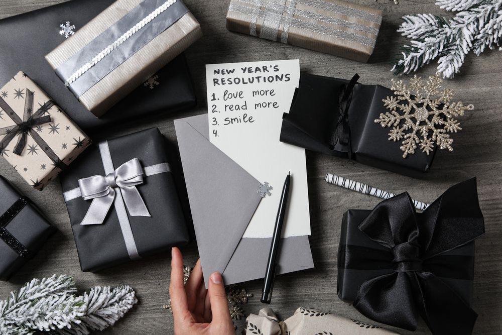 Envelope in female hand, paper with list and gift boxes on gray wooden background, top view