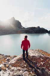 Back view of man in red jacket standing on waterside of frozen lake during daytime