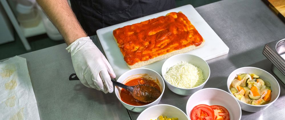 Unrecognizable male chef with gloves scooping tomato sauce to prepare pizza in restaurant kitchen