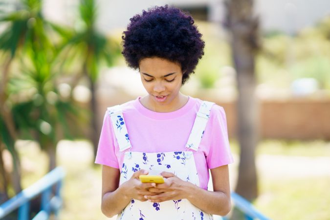 Woman in floral overalls looking down at phone outside