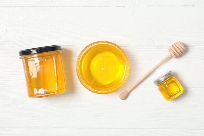 Glass jar and bowl with honey, dipper on wooden background. Flat lay