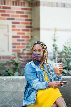 Woman wearing mask holding iced coffee and smart phone smiling and looking away