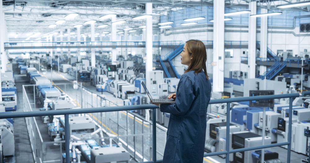 Japanese Factory Manager Working on Laptop at a Futuristic Industrial Manufacture with Autonomous AI Robots. Specialist Monitoring All the Data on a Computer, Checking Reports and Planning Research
