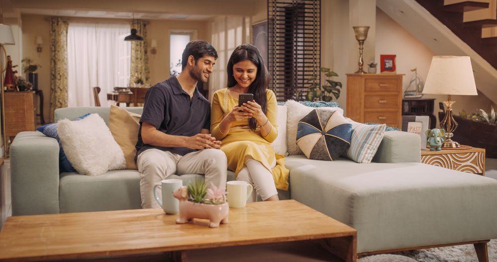 Young Indian Couple Using Internet On Smartphone, Sitting On The Sofa at Home, they Tease and Joke around.