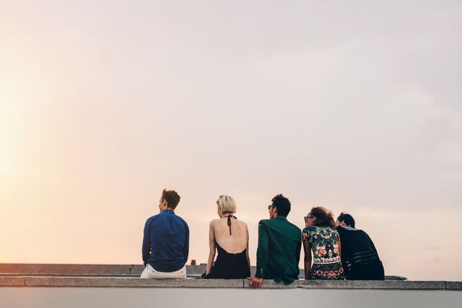 Rear view of young friends sitting together on rooftop at sunset with copy space