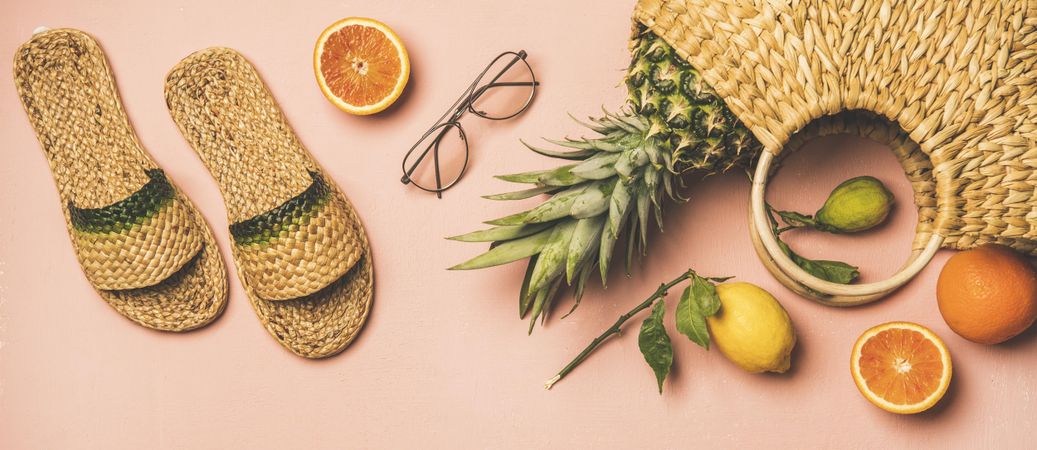 Sandals, glasses, pineapple and fruits in wicker bag, on pink background, wide composition