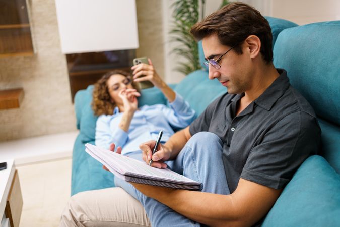 Focused husband taking notes in notebook while sitting with wife on sofa