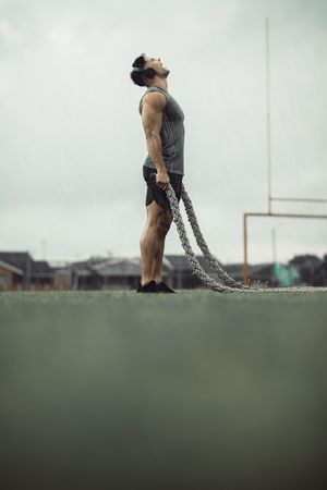 Muscular man doing fitness workout outdoors in rain with battle ropes on field