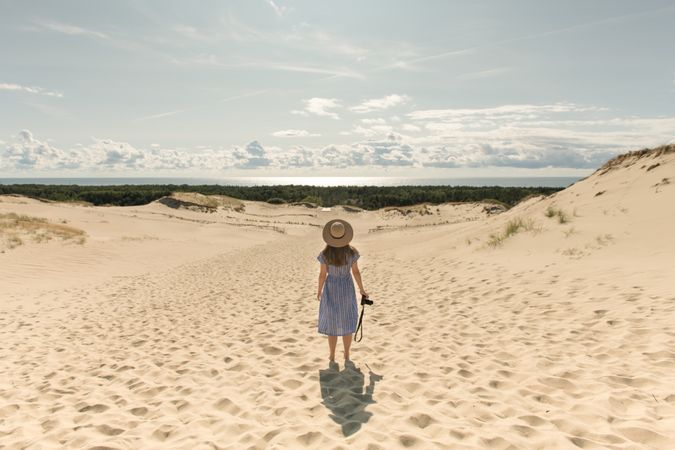 Back view of adult woman in straw hat and dress with camera walking on sand dune
