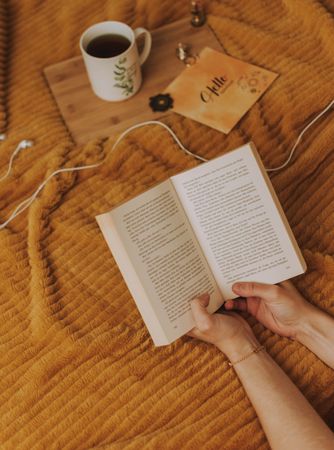 Person's hands holding a book near cup of tea on bed