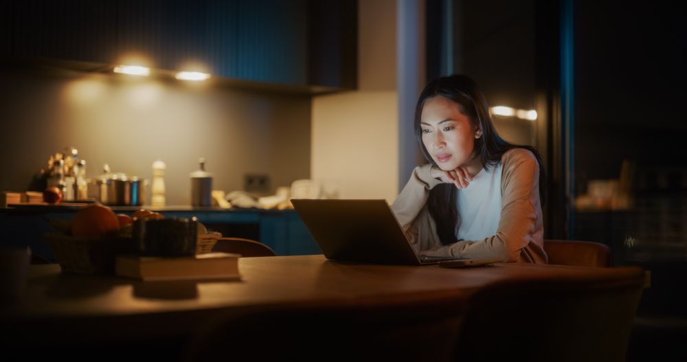 Authentic Asian Woman Sitting at a Table in a Cozy Kitchen and Using Laptop Computer at Home at Night.