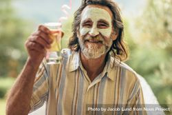 Bearded man with clay facial mask holding a glass of juice in hand bxqAX0